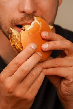 Erimiş peynirli sulu bir çizburgeri ısırmak üzere olan bir adamın yakın çekimi. Yemek ve fast food tatmini reklamları için ideal. Dikey fotoğraf.