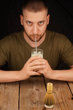 Sakallı bir adam pipetle matcha latte yudumluyor, ön planda bambu çırpısı var. Sağlıklı yaşam ve çay gelenekleri için mükemmel.