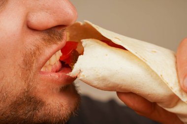 Sakallı bir adam et ve sebze rulosundan zevk alır, ağzına getirir. Resim, malzemelerin dokusunu ve tazeliğini iletir.