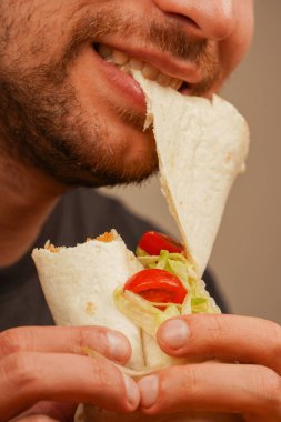 Küçük sakallı bir adam et, taze sebze ve salata dolu bir rulo pizzayı ısırır. Filmin ortasında çekilen fotoğraf, lezzetli yemekleri ve tadını vurguluyor.