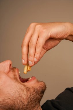 Sakallı bir adam parmaklarının arasında tuttuğu gevrek bir aperatiften bir ısırık almak için ağzını açar. Fotoğraf, kuru ekmekle atıştırmanın basit zevkini vurgular.