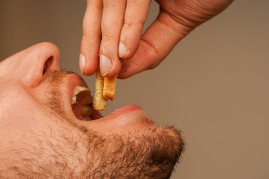 Sakallı bir adam parmaklarının arasında tuttuğu gevrek bir aperatiften bir ısırık almak için ağzını açar. Fotoğraf, kuru ekmekle atıştırmanın basit zevkini vurgular.