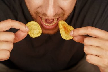 Sakallı adam altın patates cipsini ısırır. Yemek ve yaşam tarzı kavramları için mükemmel. Genç adam aynı anda iki cips yer, patates cipsinin tadını çıkarır.