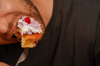 A young man in a dark T-shirt licks a cupcake skewered on a fork. A man enthusiastically eats a sweet dessert, close-up photo clipart