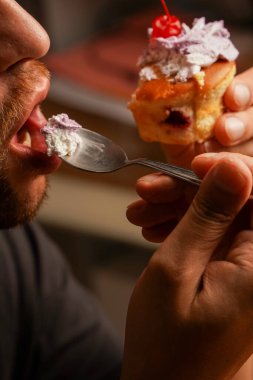 Bir adamın, elinde kiraz ve kremalı bir kaşıkla bir dilim pastanın tadını çıkardığı yakın plan. Konsept olarak, zevk içinde yaşamak, tatlı yemek ve tatlı bir şey arzulamak.