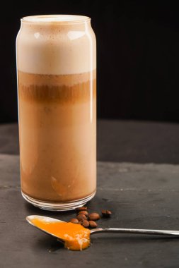 Büyük bir fincan kahve. Yanında da bir kaşık bal ve kahve çekirdeği var. Fotoğraf kalitesi iyi. Siyah bir arkaplanda