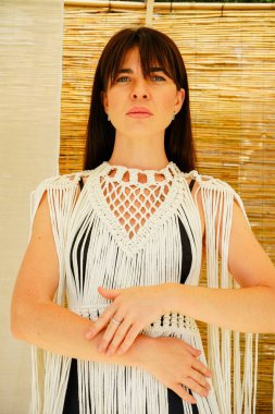 Beyaz püsküllü bir elbise giymiş, bahçesinde zarif bir şekilde duran, şifon kumaşlı ve arkasında sazdan bir duvar olan bir esmerin huzurlu görüntüsü.