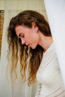 Beyaz tül arkaplanlı, narin, beyaz örülü bir elbise giymiş sarışın bir kadının yakın çekimi. Kızın kulağında gümüş bir küpe var.