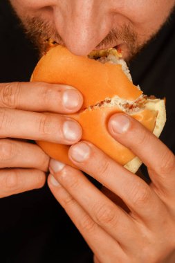 Erimiş peynirli sulu bir çizburgeri ısırmak üzere olan bir adamın yakın çekimi. Yemek ve fast food tatmini reklamları için ideal. Dikey fotoğraf.