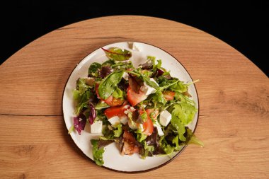 Salata ahşap bir masanın üzerinde, siyah bir arkaplanda, yakın plan fotoğraf. Sağlıklı sebze salatası, domatesli, peynirli ve tabakta otlar.