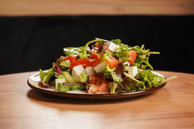 Sağlıklı sebze salatası, domatesli, peynirli ve tabakta otlar. Salata ahşap bir masanın üzerinde, siyah bir arkaplanda, yakın plan fotoğraf.