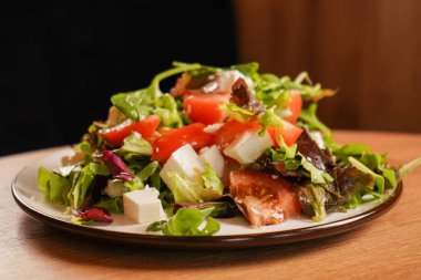 Canlı bir Yunan salatasına yakın plan, yeşil yapraklı, domatesli ve peynirli, tadını çıkarmaya hazır. Tahta masada bir tabak taze salata duruyor.