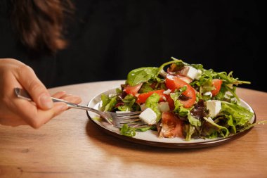 Elinde taze sebzeli, peynirli ve domatesli bir salatanın çatalı var. Tahta masadaki sağlıklı yiyecekler. Yumuşak peynirli Yunan salatası.