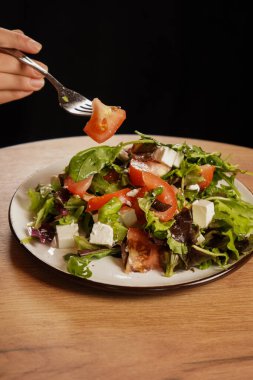 Elinde taze sebzeli, peynirli ve domatesli bir salatanın çatalı var. Tahta masadaki sağlıklı yiyecekler. Yumuşak peynirli Yunan salatası.