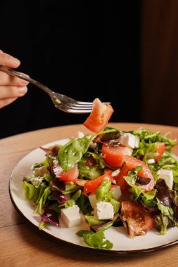 Elinde taze sebzeli, peynirli ve domatesli bir salatanın çatalı var. Tahta masadaki sağlıklı yiyecekler. Yumuşak peynirli Yunan salatası.