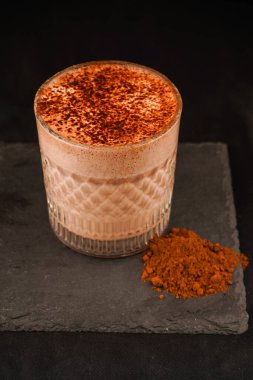 Ev yapımı kakao ya da sıcak çikolata. Cam bardağa serpilmiş çikolatayla ya da siyah arka planda kahverengi bir masada servis edilmiş fincanla süslenmiş. Bir kafe tarifi.