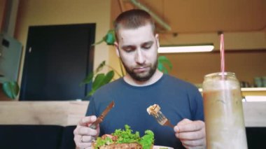 Kahverengi saçlı ve sakallı bir adamın salatalı Belçika waffle 'ı yediği rahat bir kafe sahnesi. Yeşil bitkiler ve bir içecek atmosferi tamamlar.