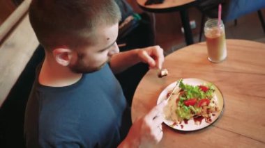 Adamın biri sıcak bir kafede mavi bir kanepeye oturmuş salatayla Belçika waffle 'ını kesiyor. Yanında pipetli bir içki, rahat bir sahne yaratıyor.