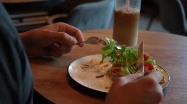 Genç bir adam yeşil bitkilerin arka planında, rahat bir kafede, ahşap bir masada Belçika waffle 'ı, salata ve domates yiyor. Masada kapaklı soğuk kahve bardağı var.