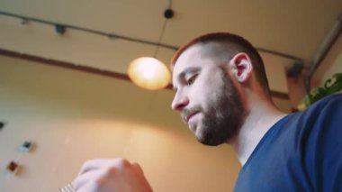 Açık kahverengi saçlı ve sakallı bir adam salata ve domatesli Belçika waffle 'ını ahşap bir masada keser. Arka planda bitkiler olan rahat bir kafe. Videonun aşağısından görüntüle
