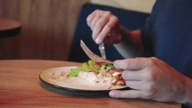 Açık kahverengi saçlı ve sakallı bir adam salata ve domatesli Belçika waffle 'ını ahşap bir masada keser. Arka planda bitkiler olan rahat bir kafe.