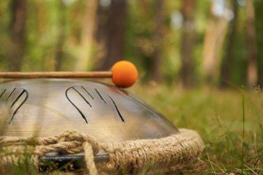 A melodic hand drum with orange-tipped sticks rests on the grass in the middle of the forest. Blurred trees create a peaceful backdrop clipart