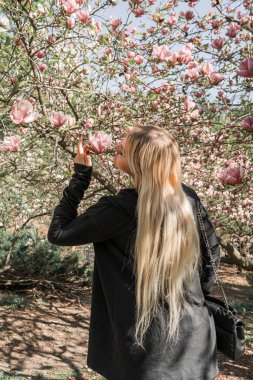 Manolya ağacının yanında baharın tadını çıkaran neşeli genç bir kadın. Parlak çiçek arkaplanı, kız çiçeğe dokunur ve kokuyu içine çeker.