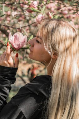 Manolya ağacının yanında baharın tadını çıkaran neşeli genç bir kadın. Parlak çiçek arkaplanı, kız çiçeğe dokunur ve kokuyu içine çeker.