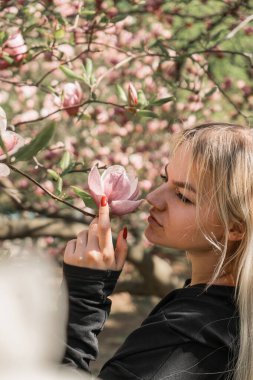 Manolya ağacının yanında baharın tadını çıkaran neşeli genç bir kadın. Parlak çiçek arkaplanı, kız çiçeğe dokunur ve kokuyu içine çeker.