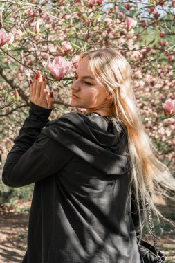 Bir kadın baharda çiçek açan bir parkın ortasında fotoğraf için poz veriyor. Arka planda pembe manolya çiçekleri var. Siyah ceketli, beyaz saçlı bir kadın.