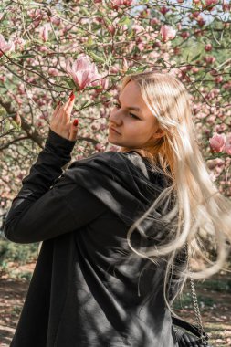 Siyah elbiseli ve uzun beyaz saçlı bir kız manolya ağacındaki çiçeklere uzanır. Çerçevesinde çiçekler olan bahar atmosferi