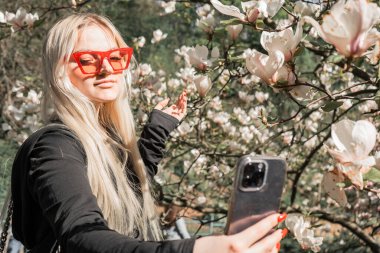 Manolya çiçekleriyle çevrili gözlüklü bir kadın selfie çekiyor. Bahar cazibesi ve çiçek açan odak noktası, telefonda selfie.