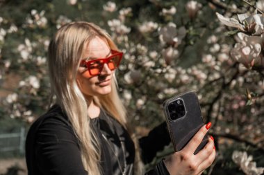 Genç sarışın kız baharda telefonuyla selfie çekiyor. Çiçek açan bir manolya ağacının arka planında, gözlüklü bir kadın çerçevesinde selfie çekiyor, büyük çiçek tomurcukları.