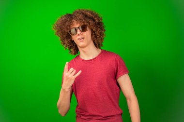 Kıvırcık saçlı ve şık gözlükleri olan genç bir çocuk yeşil krom anahtar arka planda gelişigüzel duruyor. Dengeli tavrı bu uyumlu görsel kompozisyona cazibe ve karmaşıklık katıyor.