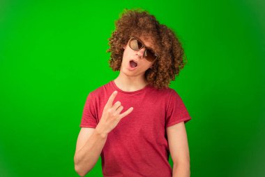 Kıvırcık saçlı çocuk, canlı bir yeşil krom anahtar zeminine karşı, gözlüklerini düşünceli bir şekilde ayarlıyor, çeşitli yaratıcı uygulamalar için profesyonel ama yaklaşılabilir bir görünüm yaratıyor.