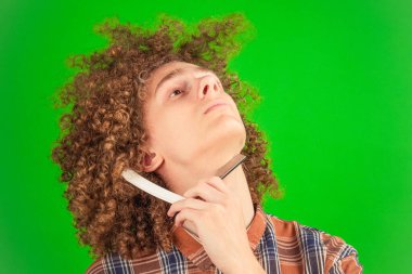 Canlı bir yeşil krom anahtara karşı, resmi tişört giymiş kıvırcık saçlı bir çocuk yüzünü nazikçe tıraş ediyor, yaratıcı multimedya projeleri için uyumlu ve profesyonel bir sahne sağlıyor.