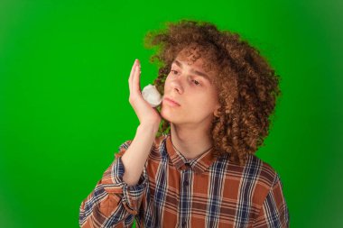 Dikkatle köpüren, kıvırcık saçlı, temiz gömlekli çocuk, dinamik ve profesyonel bir görsel görüntü için yeşil krom anahtar zemin üzerinde durarak yüzüne sürtünüyor.