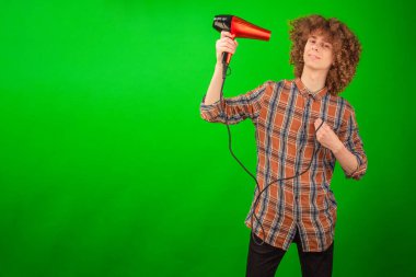 Hareket halindeyken yakalanan kıvırcık saçlı bir adam saç kurutma makinesinin yüzüne üflemesine izin verir. Temiz gömleği yeşil bir arka planda dalgalanır.