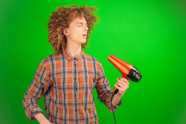 Yapay rüzgarla yıkanan, gömlekli kıvırcık saçlı adam, parlak yeşil bir arka planla çerçevelenmiş bir saç kurutma makinesiyle teatral poz veriyor.