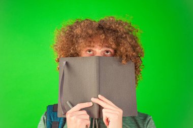 Kendinden emin ve sakin olan kıvırcık saçlı adam, yeşil gömlekli, kalın Chroma Key arka planına karşı bir defter tutuyor.
