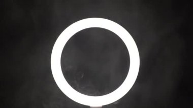 A stream of smoke in front of a ring of light from a led lamp on a black background close-up. The vaper blows steam in the frame. The concept of a dance club and a stage with spotlights.