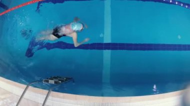 Eşofmanlı, dalış gözlüklü, mayo takan kadın yüzücü havuzda antrenman yapıyor. Sporcu kelebek stilinde yüzer. Geniş açı, 4k..