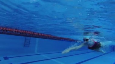 Eşofmanlı, dalış gözlüklü, mayo takan kadın yüzücü havuzda antrenman yapıyor. Sporcu su altında yüzer. Sualtı çekimi, geniş açı, 4K.