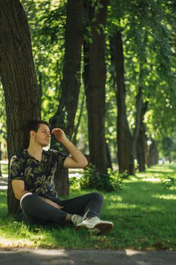 Genç bir adam parkta bir ağacın altında oturur, bagaja yaslanır. Rahat duruşu ve koyu renk elbisesi sakin ve huzurlu bir atmosfer yaratıyor. Park ayarı sükuneti artırıyor