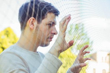 Haşin bir ifadeyle genç bir adam örgüden dışarı bakar. Bakışlarının ciddiyeti derin bir tefekkür hissi veriyor. Onun özellikleri yoğun duyguların özünü yakalar.