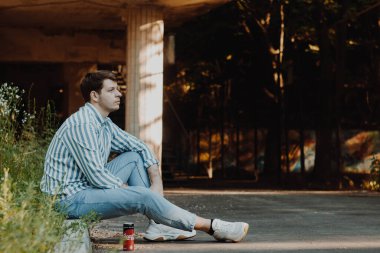 Kaldırım kenarına oturmuş, açık renkli günlük kıyafetler giyen bir adam derin bir ifadeyle mesafeye bakıyor. Ayarlama, dinginliği ve derin duygusal derinliği iletir.