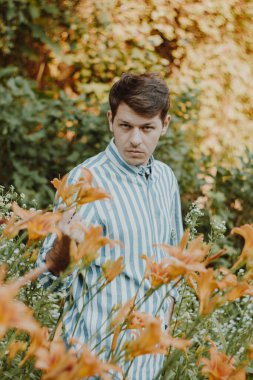 Çizgili tişörtlü bir adam çiçek açan bir bahçede zambaklarla çevrili duruyor. Barışçıl ortam, doğayla uyum ve bağın bir yansımasıdır. Gerçekten huzurlu bir an.
