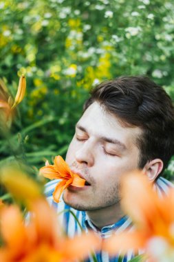 Koyu renk saçlı Avrupalı bir adam ağzında turuncu bir zambak tutuyor. Ağaçların ve yeşil bitkilerin doğal arka planı doğayla bir uyum duygusu yaratır. Görüntü barışçıl enerji yayıyor.