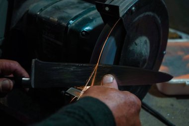 Bir zanaatkârın eli, öğütme çarkında metal bir aleti bilerken yakın plan çekilmiş. Sürtünme parlak kıvılcımlar yaratır. Atölyenin loş arka planı sanayi sürecini vurguluyor