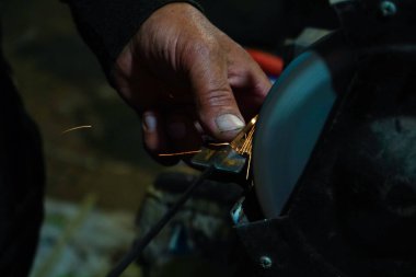 Bir zanaatkârın eli, öğütme çarkında metal bir aleti bilerken yakın plan çekilmiş. Sürtünme parlak kıvılcımlar yaratır. Atölyenin loş arka planı sanayi sürecini vurguluyor